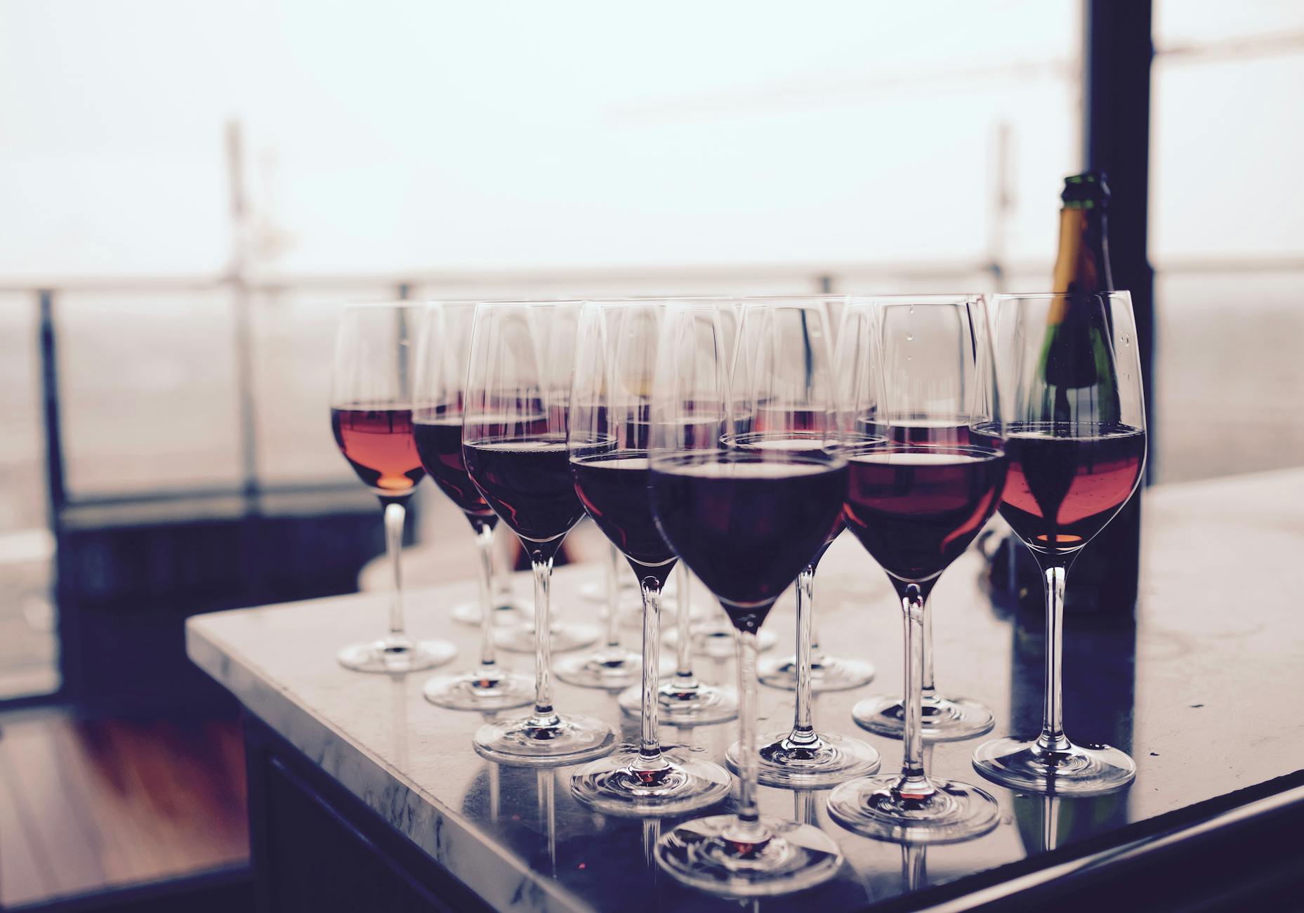 Line of wine glasses filled with different types of red wine, arranged on a tabletop with a bottle in the background.