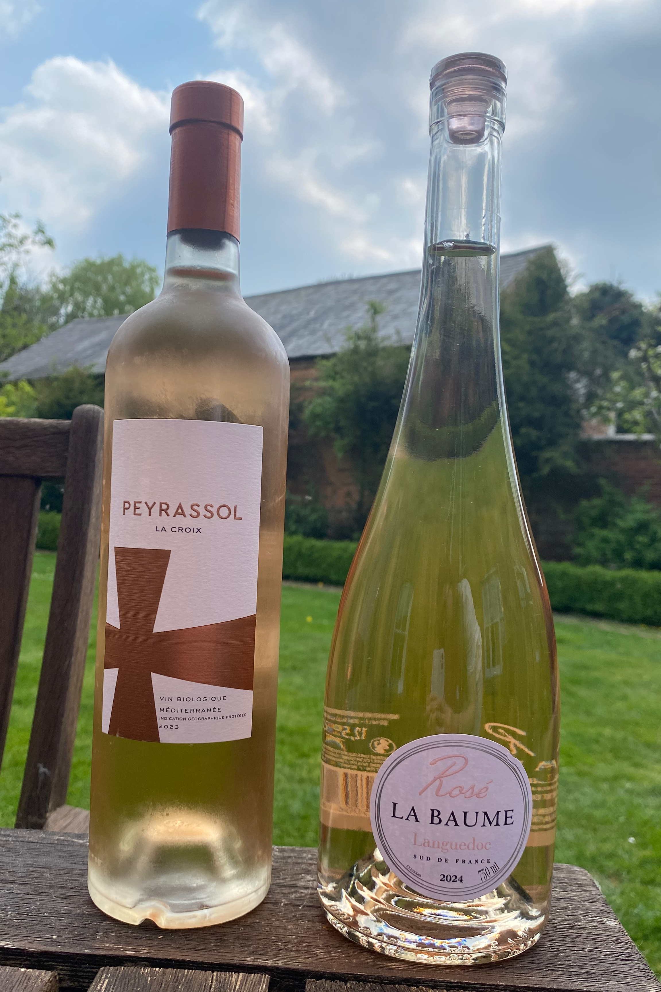 Two bottles of rosé wine on a wooden table, with a blurred garden background. The left bottle is labeled 'Peyrassol La Croix,' while the right bottle is labeled 'Rosé La Baume Languedoc.'