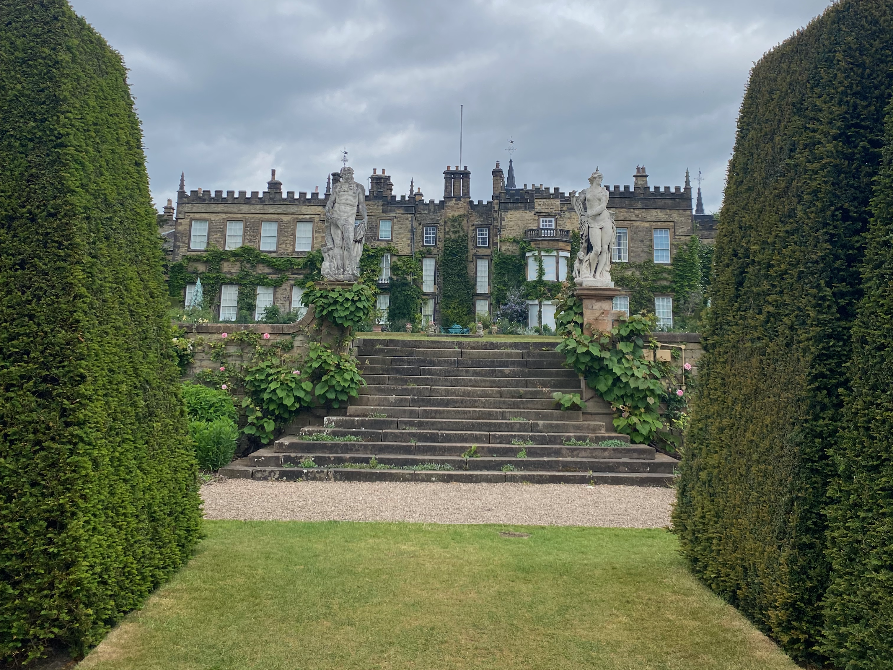 A view of a grand estate with statues flanking the stairs leading up to the building, surrounded by neatly trimmed hedges and overcast skies.