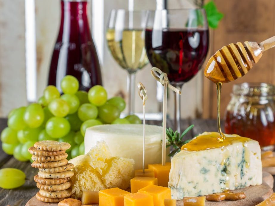 A selection of cheeses including blue cheese and crackers served with grapes, honey drizzled over cheese, and glasses of red and white wine in the background.