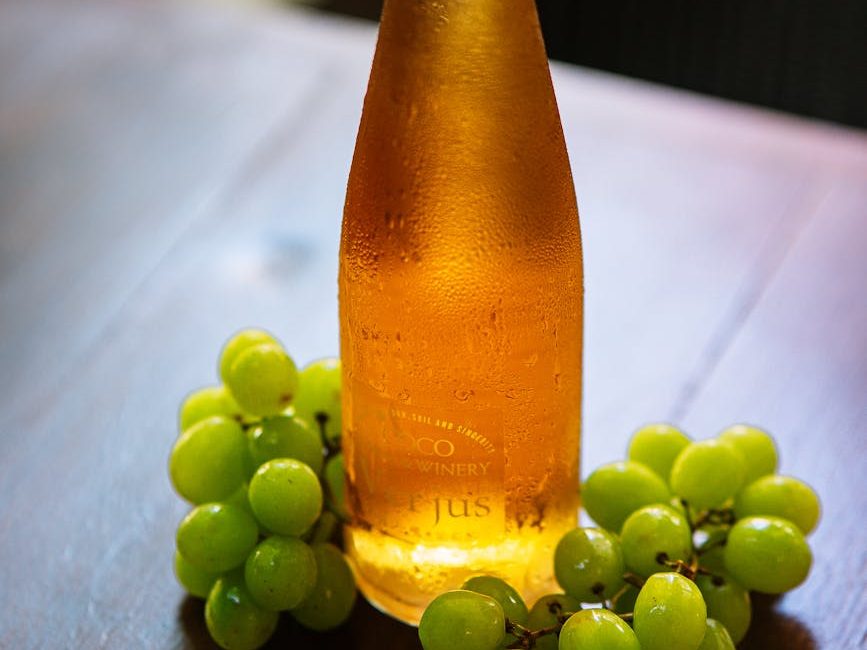 A bottle of amber-colored orange wine next to clusters of green grapes on a wooden table.