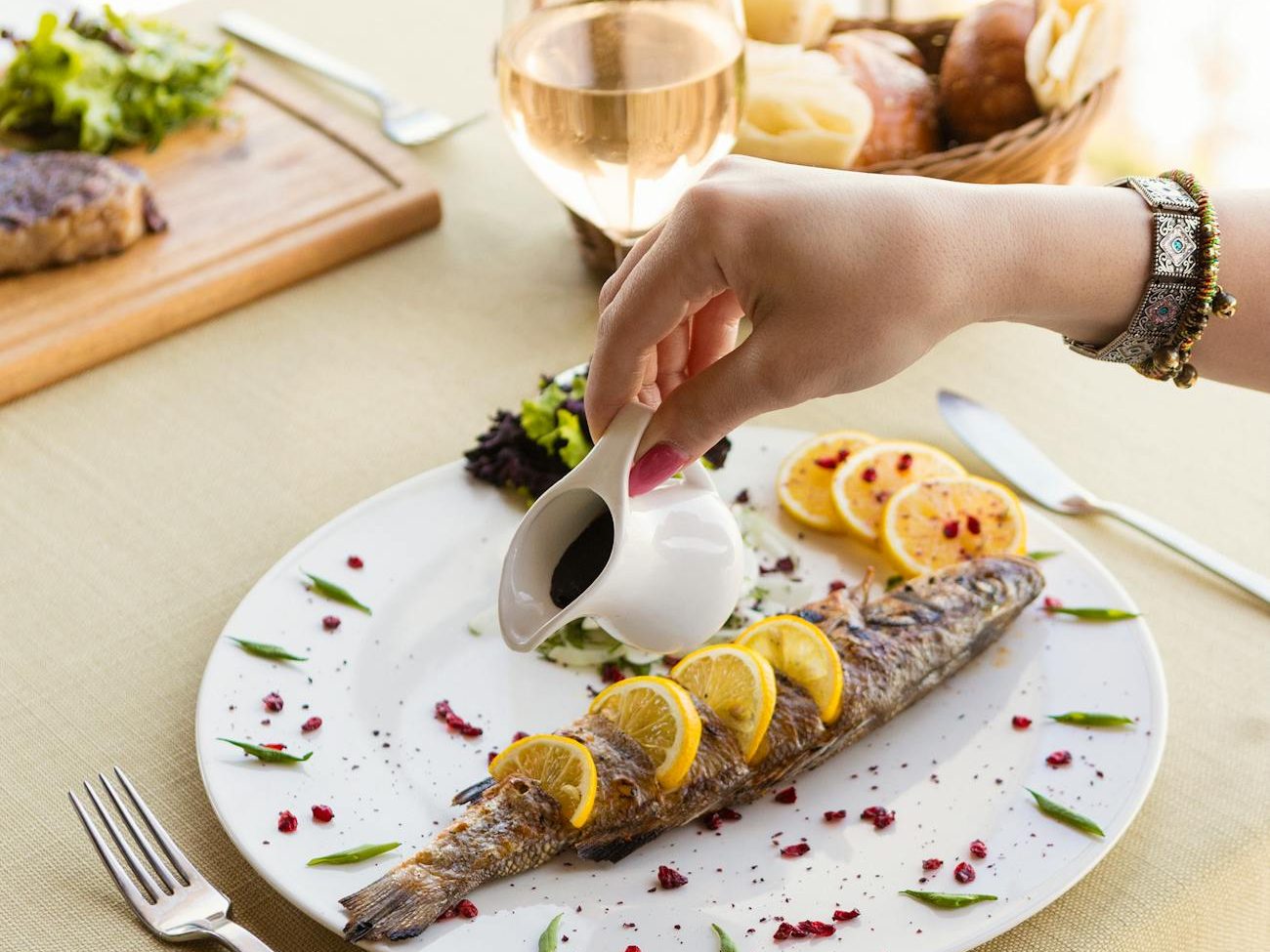 A hand pours sauce over a beautifully plated oily fish dish, garnished with lemon slices and fresh greens, accompanied by a glass of white wine.