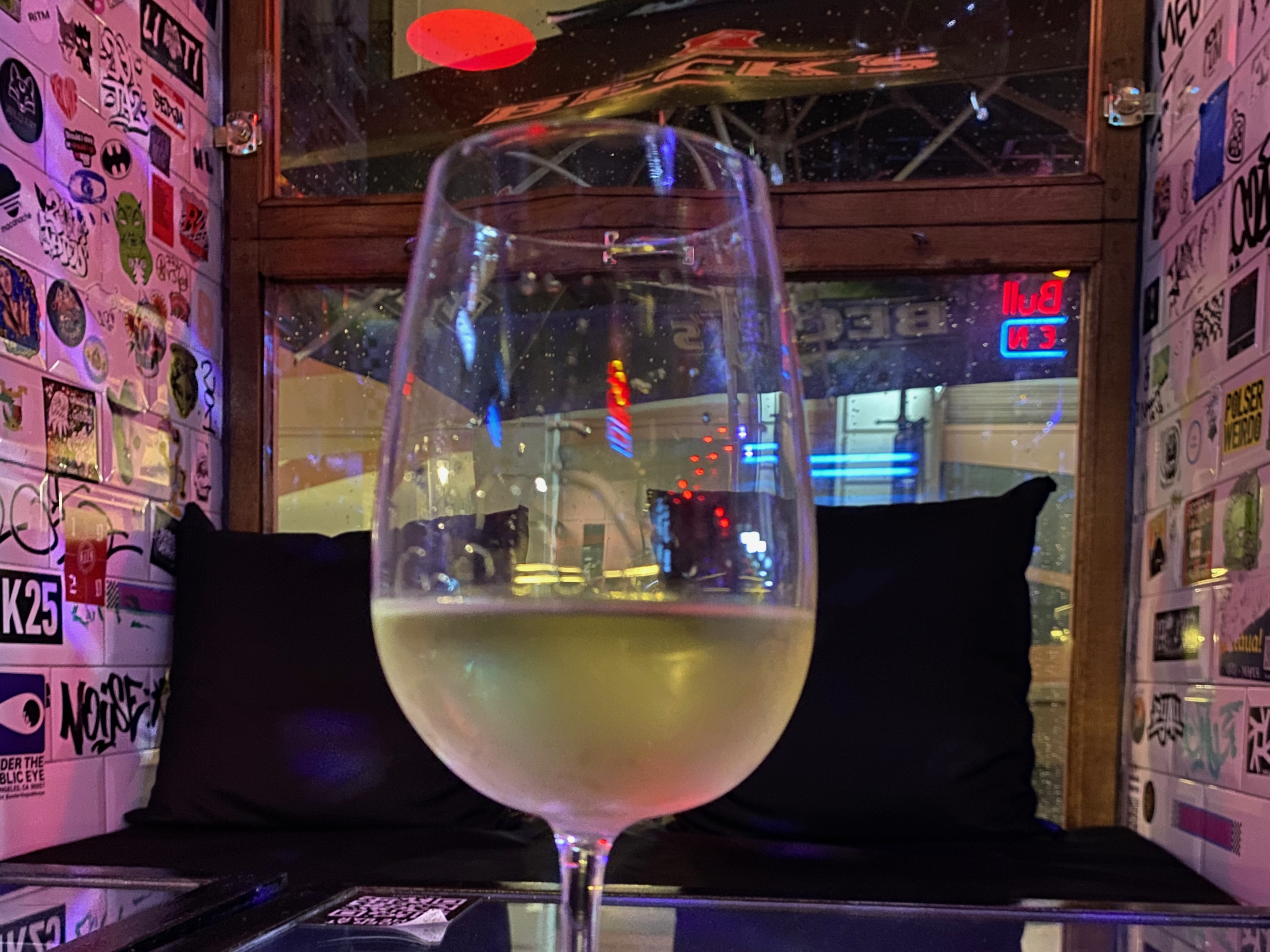 A glass of white wine sits on a table in a cozy bar, surrounded by colorful and eclectic stickers on the walls.