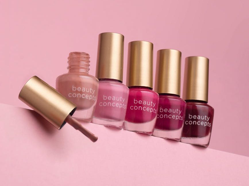 A collection of beauty concept nail polish bottles in various colors, arranged on a pink background, with one bottle opened and its brush extended.