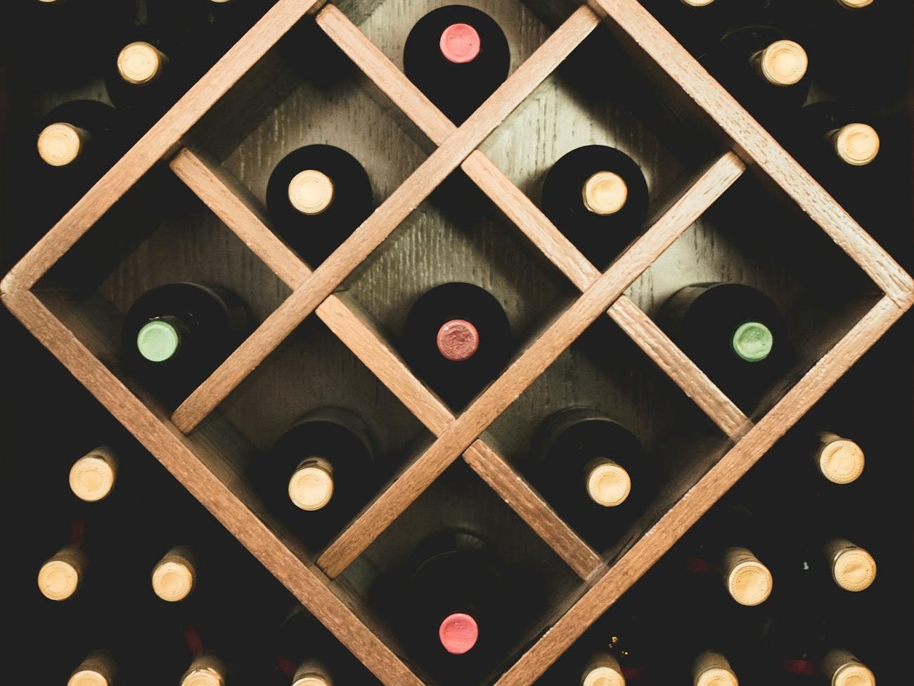 A wooden wine rack displaying various wine bottles with colored caps arranged in a diamond pattern.