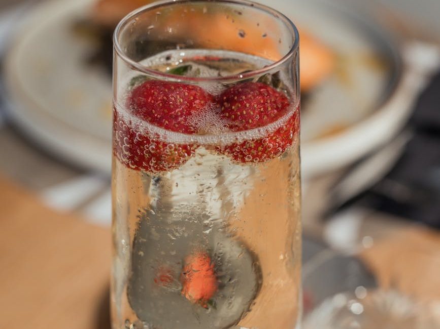 A glass of sparkling wine with strawberries floating on top, set against a blurred background featuring a plate of food.