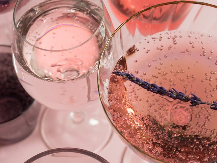 A close-up of sparkling rosé wine in a glass with bubbles and a sprig of lavender, surrounded by other glasses of pink and clear beverages.