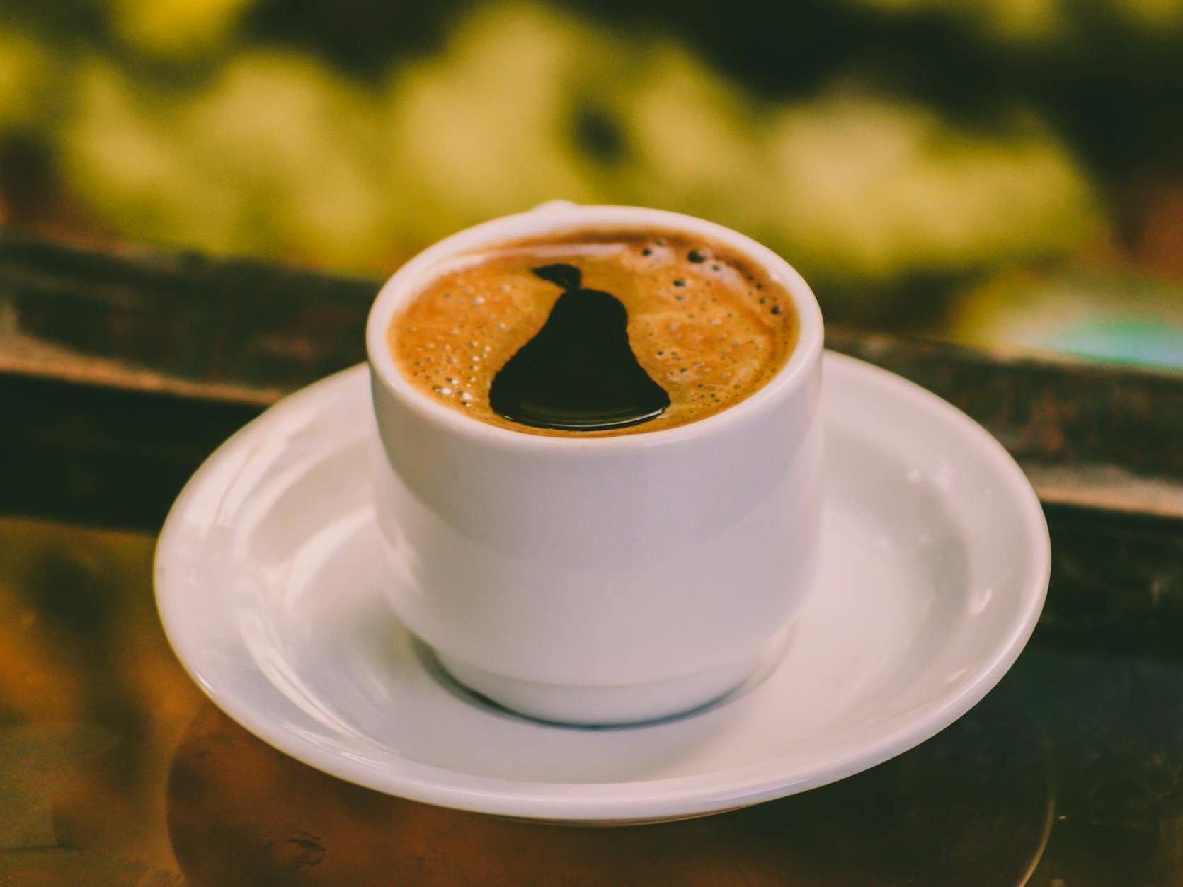 A close-up image of a coffee cup with rich, dark espresso sitting on a white saucer, set against a blurred background.
