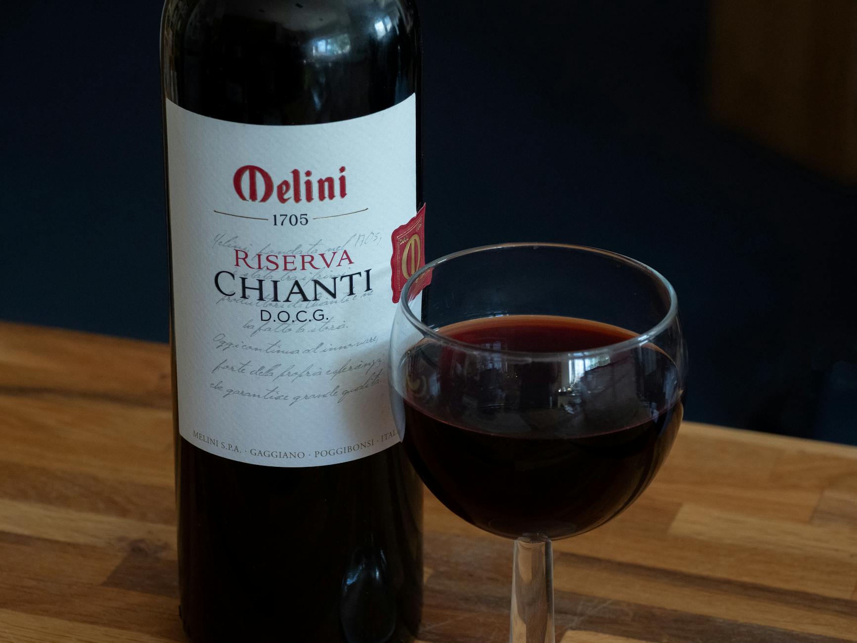A bottle of Melini Riserva Chianti D.O.C.G. alongside a glass filled with dark red wine, placed on a wooden surface.