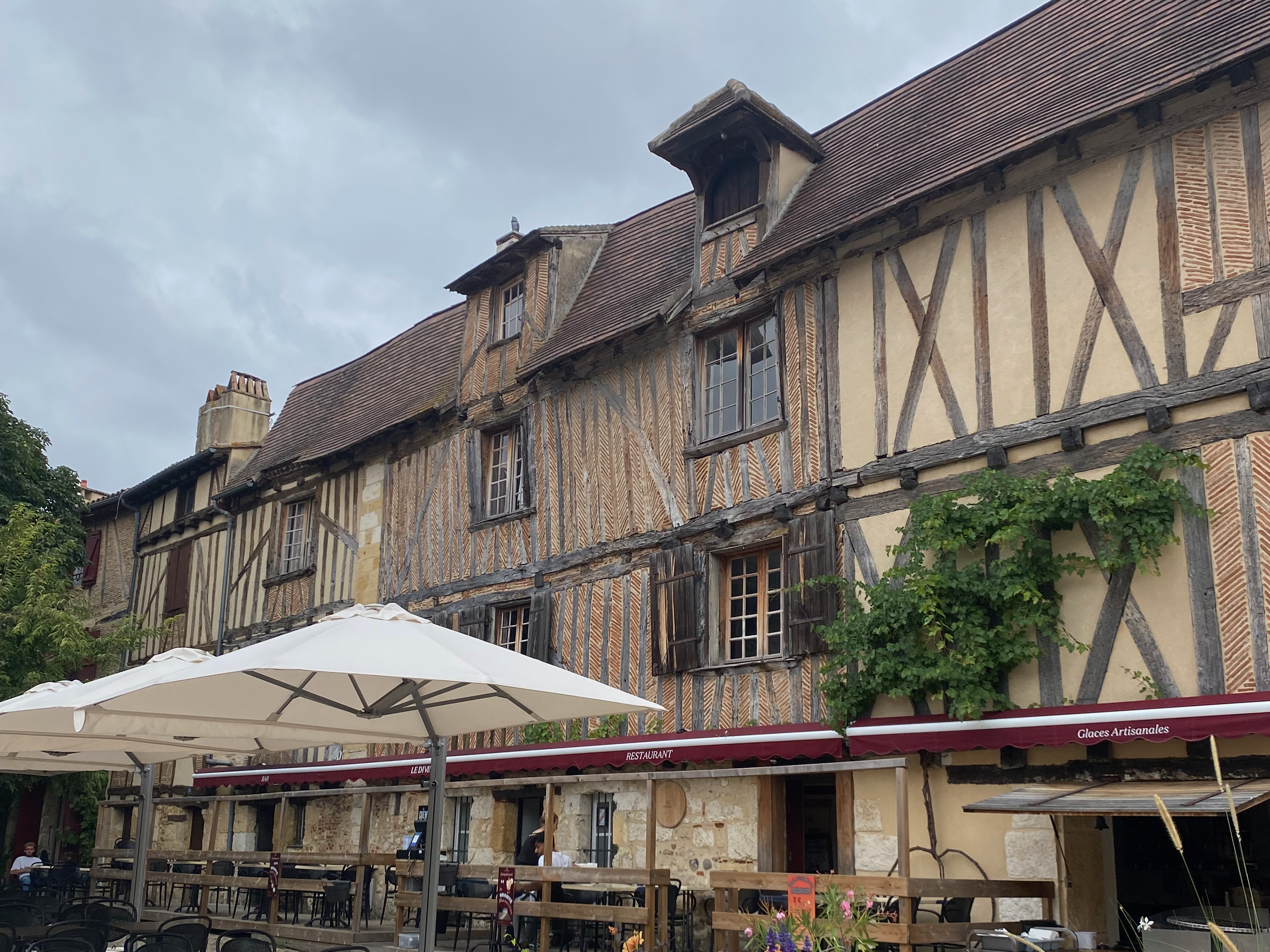 A traditional timber-framed building with a restaurant, featuring flower beds with colorful blooms and outdoor seating under umbrellas.